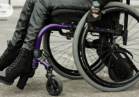 jambes d'une femme avec chaussures à talons et un pantalon de type jeans bleu qui est sur un fauteuil roulant manuel violet en train de faire le geste de rouler en ayant ses mains posées sur les roues.