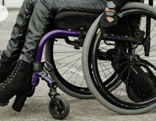 jambes d'une femme avec chaussures à talons et un pantalon de type jeans bleu qui est sur un fauteuil roulant manuel violet en train de faire le geste de rouler en ayant ses mains posées sur les roues.