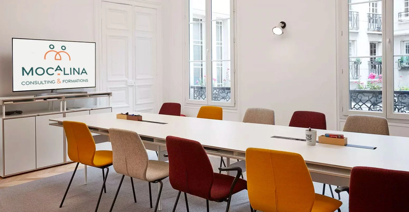 Salle de formation avec une table large et des chaises de chaque côté. Un écran de télévision affiche le logo de Mocalina, consulting et formations